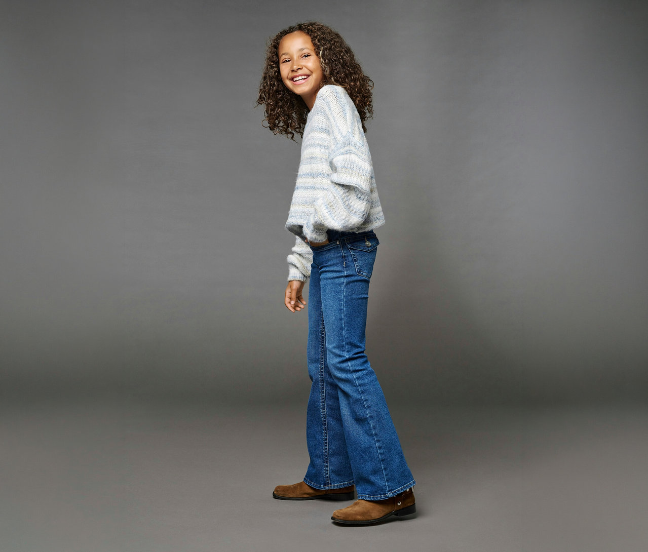 Lächelndes junges Mädchen mit lockigem Haar, das über die Schulter blickt und einen hellblau-weiß gestreiften Pullover, blaue ONLY GIRLS Phoebe Flared-Jeans und braune Schuhe trägt.