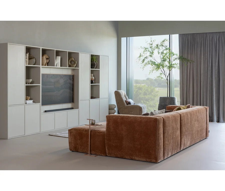 Ein modernes Wohnzimmer mit einem großen, strukturierten braunen L-förmigen Sofa, einer hellgrauen modularen Wohnwand mit Fernseher und Dekoartikeln sowie einem beigen Sessel an einem großen Fenster mit Blick auf eine grüne Landschaft. Ein kleiner Topfbaum steht neben dem Fenster.