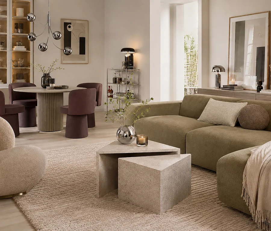 Ein modernes offenes Wohn- und Esszimmer. Der Wohnbereich hat ein olivgrünes Modulsofa, einen beigen Teppich und zwei ineinander verschachtelte dreieckige Steintische mit einer silbernen Vase und Kerze. Der Essbereich hat einen runden Tisch mit geriffelter Basis, vier violetten Samtstühlen und einen silbernen Kugelleuchter. Eine Bar und eine Vitrine sind im Hintergrund zu sehen.