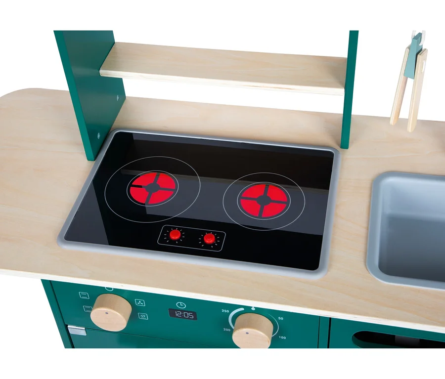 Nahaufnahme einer Spielküche mit heller Holzplatte und dunkelgrünem Unterbau. Sie verfügt über ein schwarzes Kochfeld mit zwei roten Kochplatten, ein graues Spülbecken und hölzerne Bedienknöpfe mit einer Digitalanzeige, die 12:05 anzeigt. Ein Holzregal und eine Zange sind ebenfalls zu sehen.
