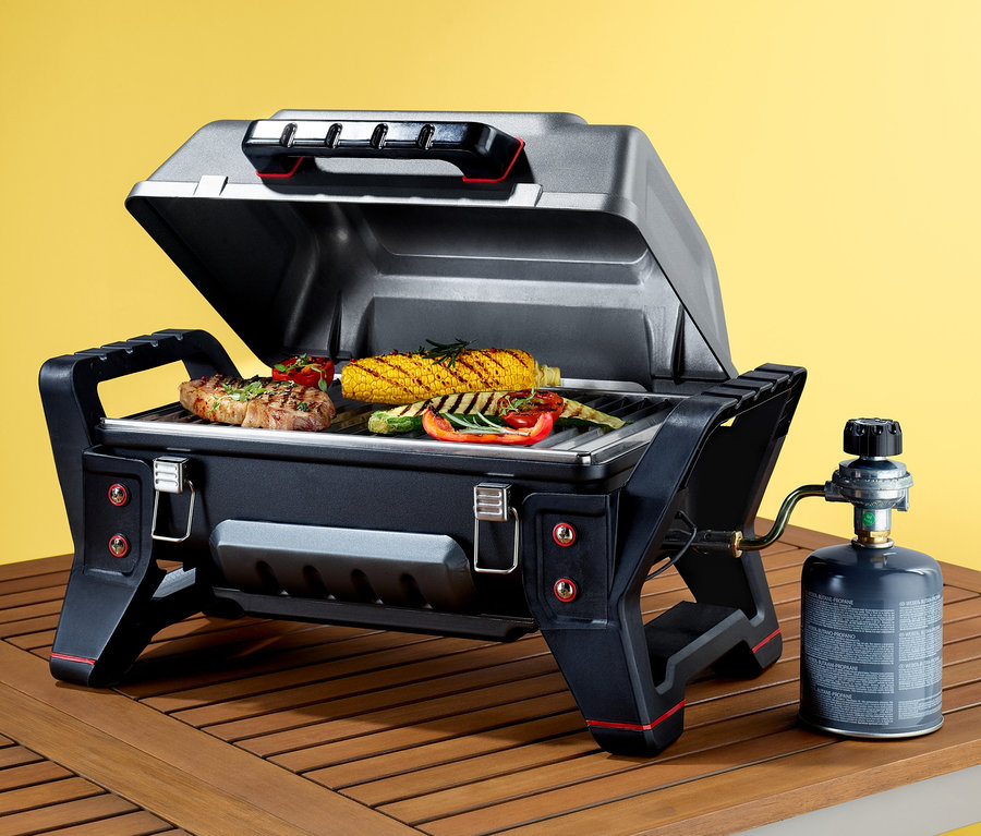 Auf einem Holztisch steht ein tragbarer Gasgrill Char-Broil® Grill2Go mit geöffnetem Deckel und Speisen im Inneren.