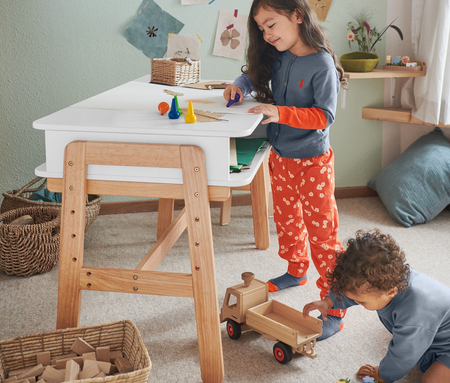 Zwei Kinder spielen in einem Kinderzimmer. Ein Mädchen zeichnet mit 5 Wachsmalstiften am Tisch. Ein Junge spielt am Boden mit Bauklötzen und trägt Baby-Sweat-Strampler und eine Kleinkind-Schlupfhose mit Blumen-Alloverprint.