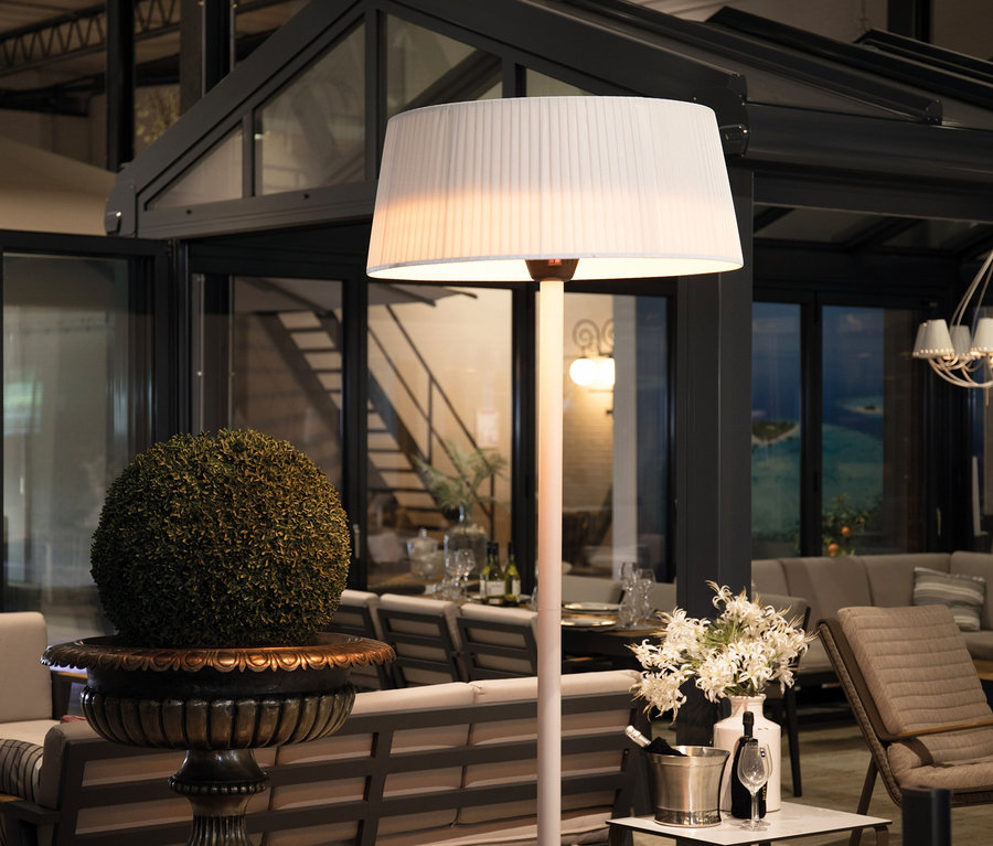 Eine Stehlampe mit weißem Schirm steht in einem Wohnbereich. Im Hintergrund ist der Sunred-Stand-Heizstrahler »Halogen Artix Basic« zu sehen.