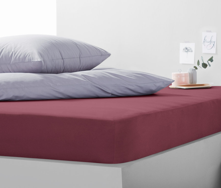 Weinrotes Jersey-Spannbettlaken in Doppelgröße mit grauen Kissen auf einem Bett.