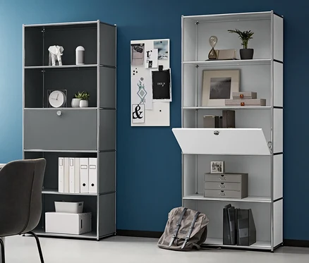 Zimmer mit Schreibtisch, Stuhl und zwei hohen Metallregalen mit einem versetzbaren Klappenfach. Links steht ein Metall-Rollcontainer, auf dem Tisch ein Laptop.