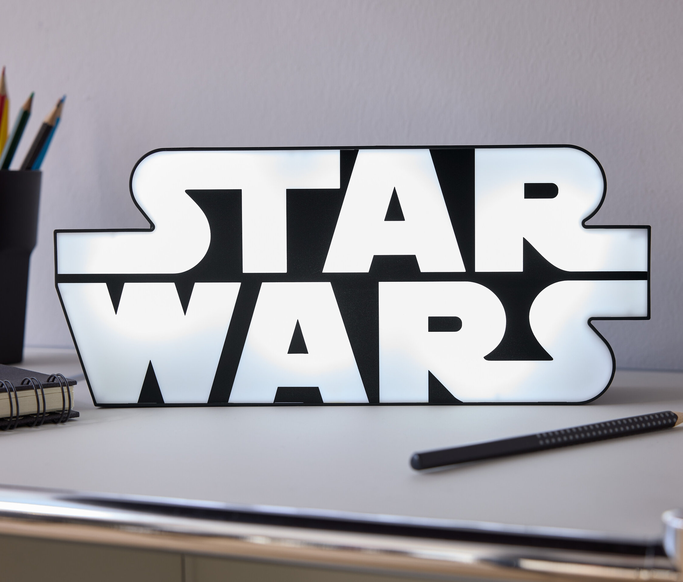 Auf einem Tisch stehen ein leuchtendes Star Wars-Schild, ein Bleistift, ein Notizbuch und ein Becher mit Buntstiften.