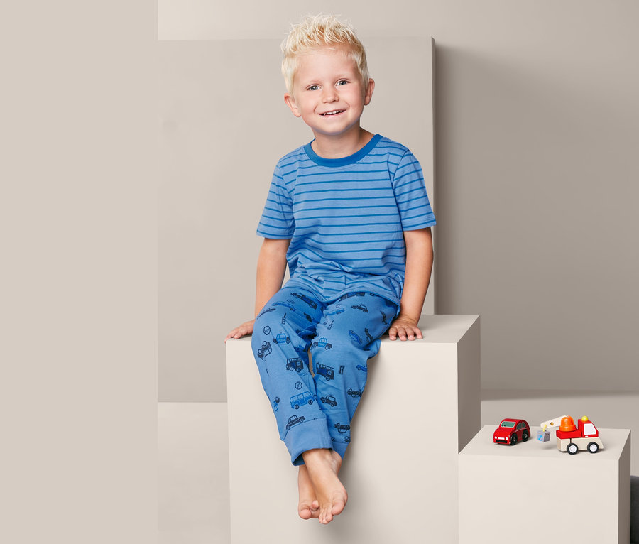 Ein Junge sitzt auf einem weißen Würfel und trägt einen blauen 3-teiligen Pyjama mit Autoaufdruck.