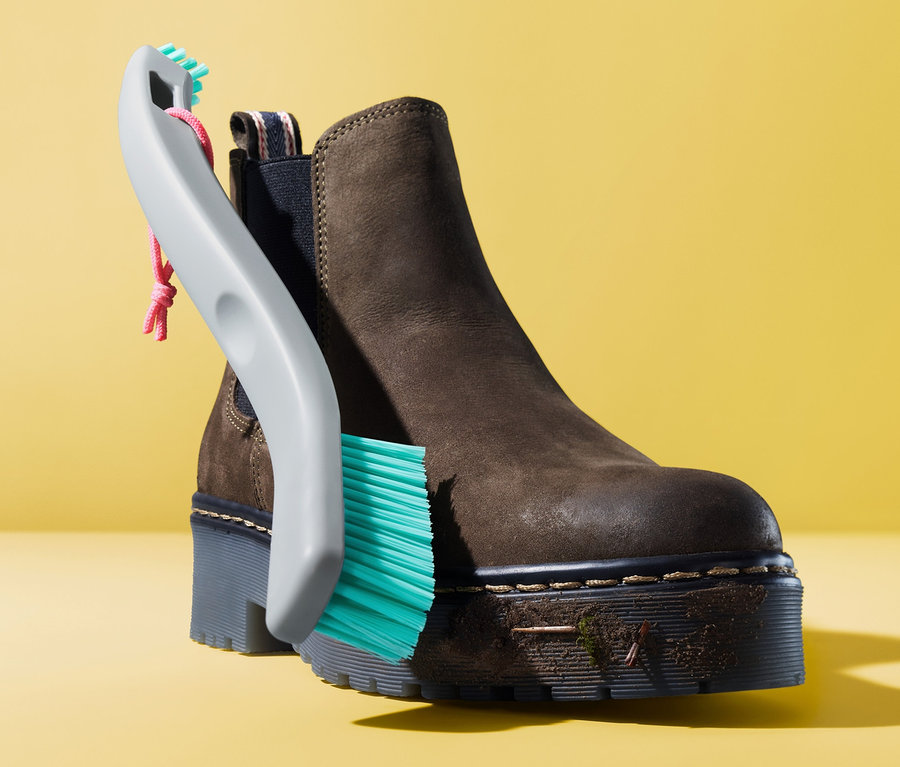 Ein brauner Stiefelette wird mit einer 2-in-1-Schuhreinigungsbürste vor einem gelben Hintergrund gereinigt.