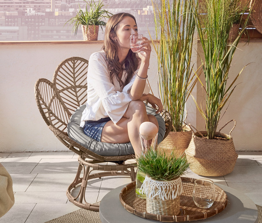 Frau sitzt auf einem Drehkorbsessel »Flower« und trinkt Wasser. Im Hintergrund befinden sich eine Gartenbank und ein Loungesessel »Boho«.