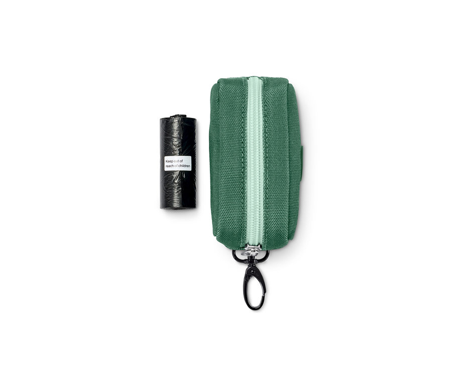 Eine grüne Hunde-Gassibeutel-Tasche mit silbernem Reißverschluss und schwarzem Karabiner neben einer schwarzen Rolle Kotbeutel.