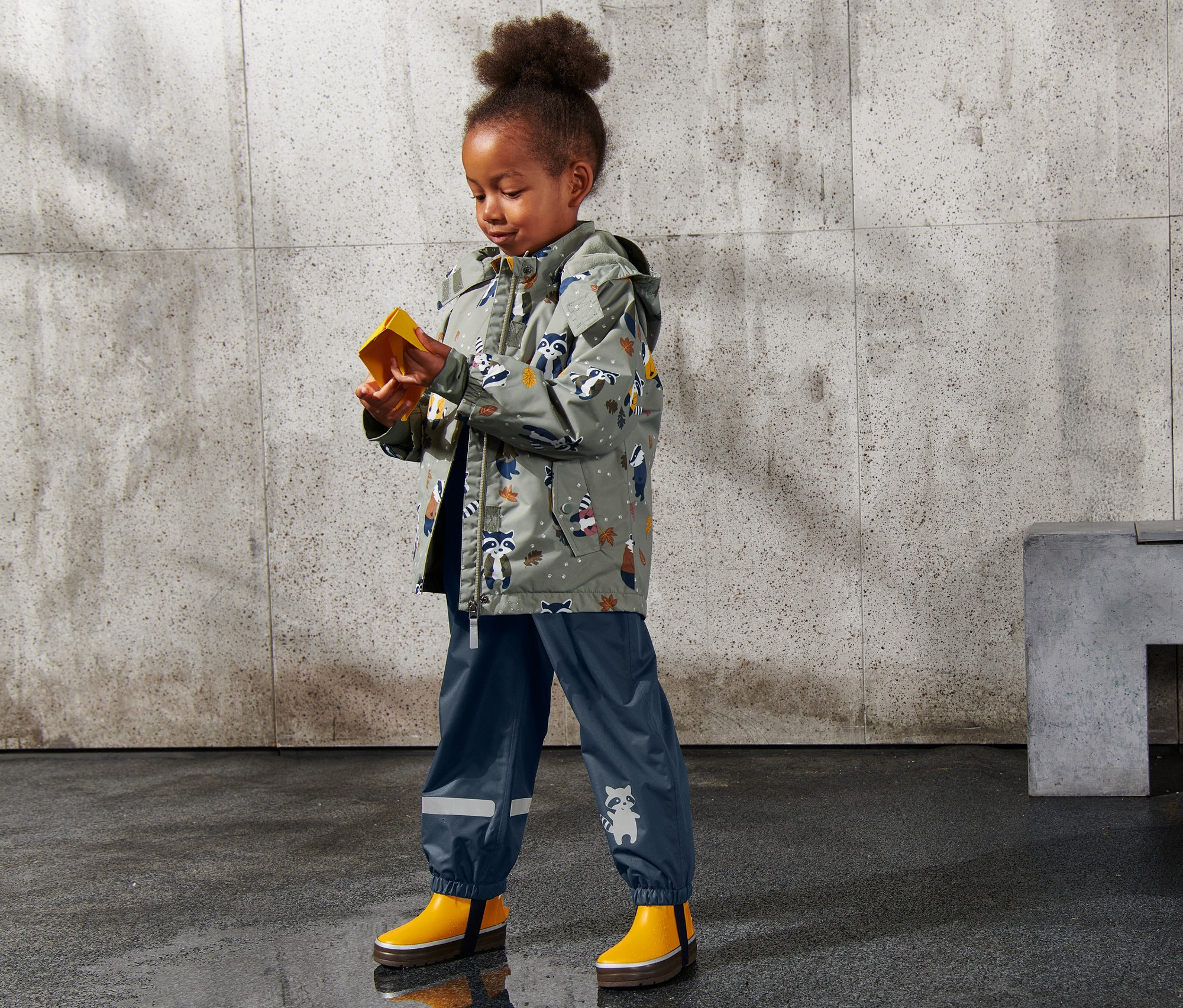 Ein kleines Mädchen untersucht eine gelbe Schachtel auf einer nassen Oberfläche. Sie trägt eine Jacke mit Waschbär-Muster und eine blaue Regenhose.