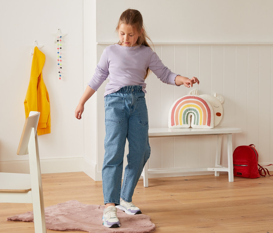 Mädchen steht auf einem rosa Teppich und trägt eine High-Waist-Jeans.