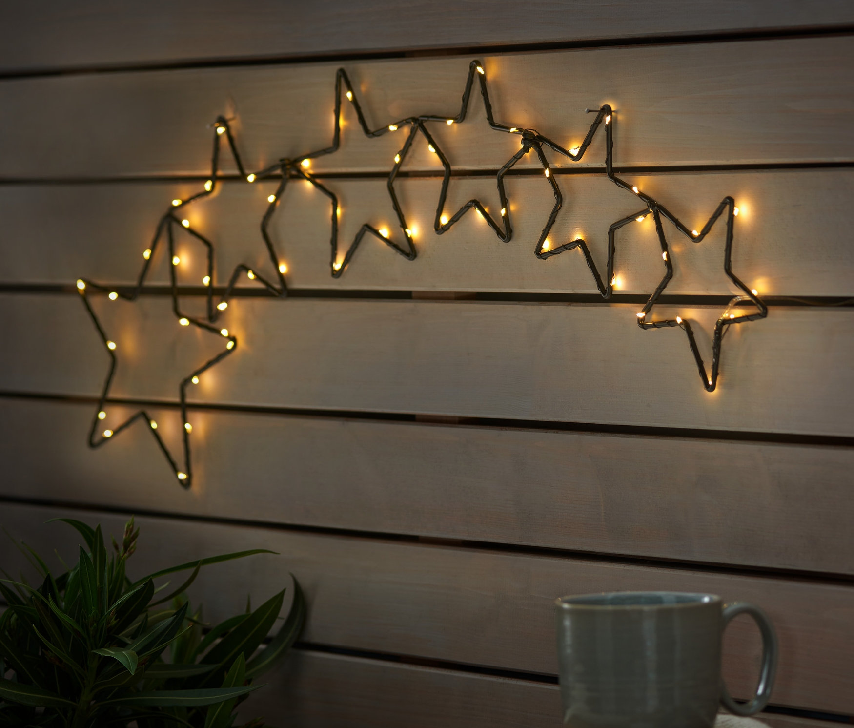 An einer Holzwand hängen LED-Outdoor-Sternensilhouetten. Auf einem Holztisch stehen eine Tasse und ein Teller mit Sternen.