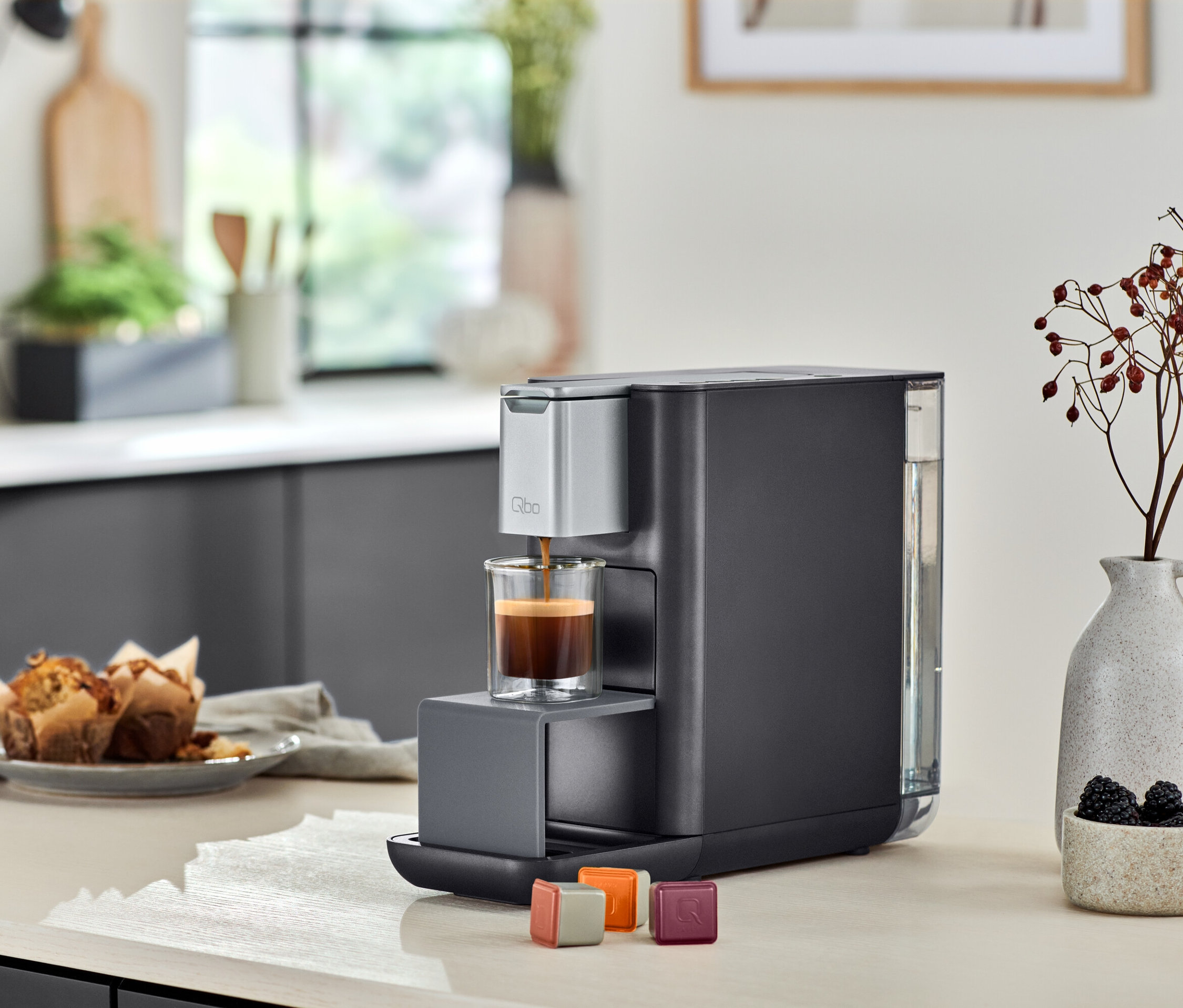 Qbo ESSENTIAL Dark Stone Kaffeemaschine füllt ein Glas mit Kaffee. Daneben stehen Qbo ICONIC.