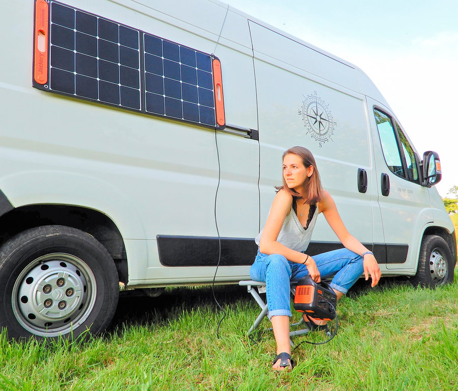 Frau sitzt auf einem Hocker neben einem weißen Lieferwagen mit Jackery SolarSaga 100 Panel und hält Jackery Explorer 240.