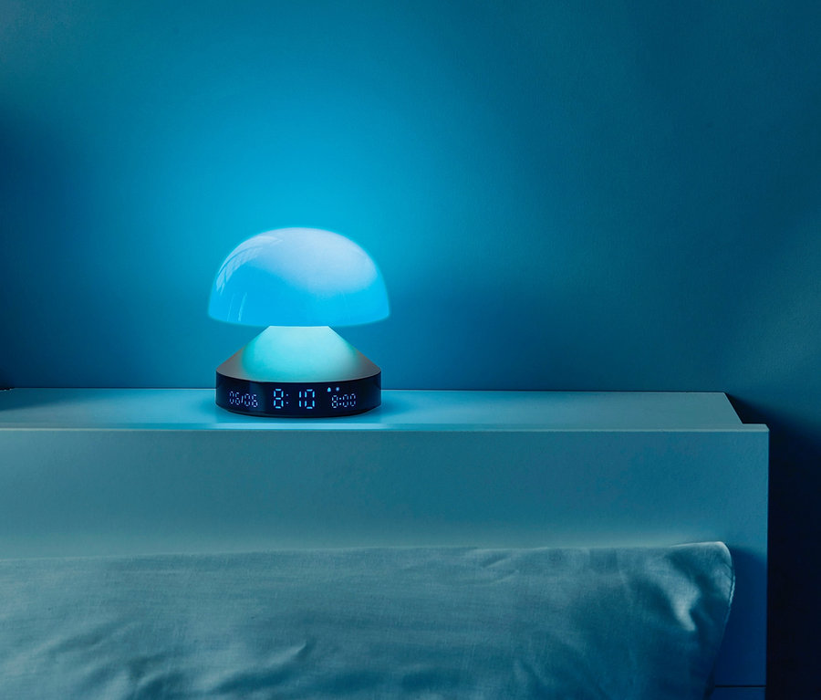 Nachttisch mit Büchern, einem grauen Blumentopf und einem anthrazitfarbenen LEXON Lichtwecker »Mina Sunrise«.