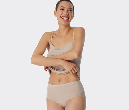 Eine lächelnde Frau hält den Saum ihres Tanktops. Sie trägt sandfarbene SCHIESSER Midislips mit Streifen.