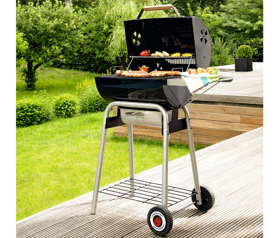 Ein schwarzer Landmann Holzkohlegrill »Black Taurus 660« mit Grillgut steht auf einer Holzterrasse.