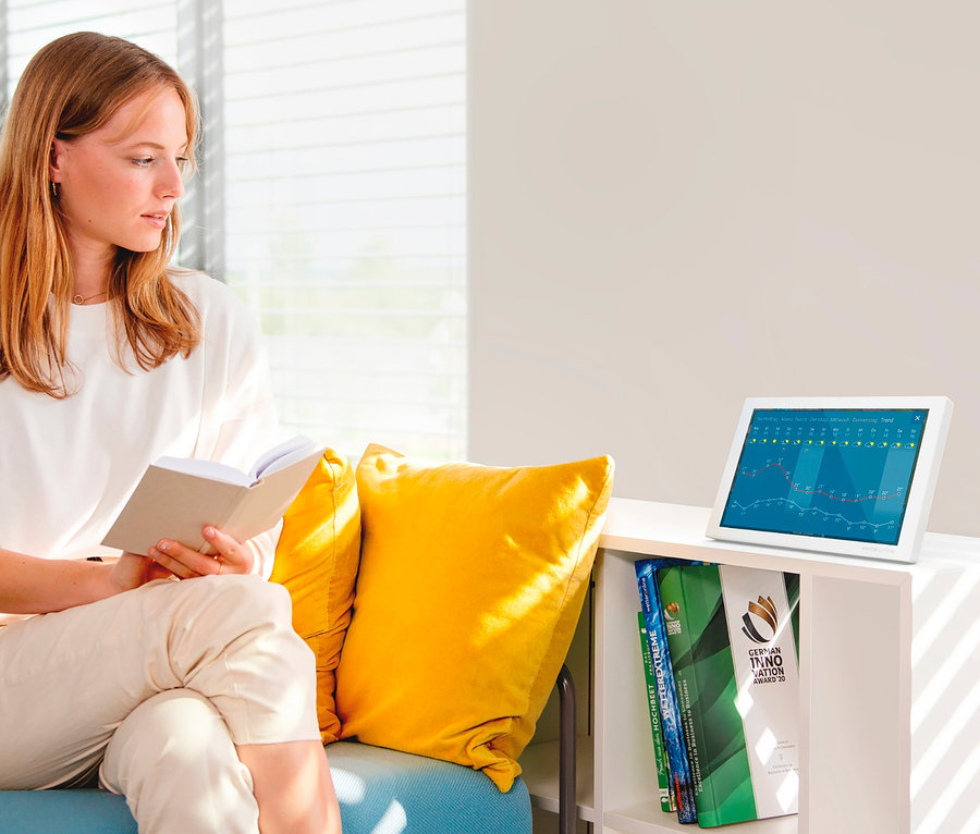 Frau liest ein Buch, während sie auf einem blauen Sofa neben einem gelben Kissen und einem weißen WetterOnline WLAN-Wetter Display Home 3 sitzt.