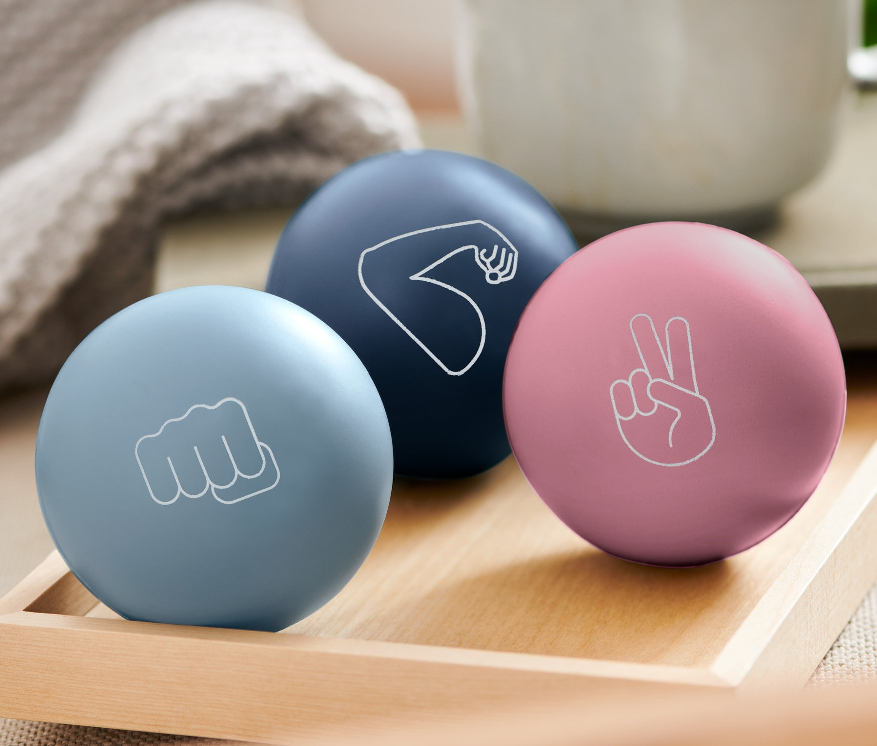 Drei Anti-Stressbälle liegen auf einem Holztablett: hellblau mit einer Faust, dunkelblau mit einem Arm und rosa mit einem Friedenszeichen. Im Hintergrund eine Tasse.