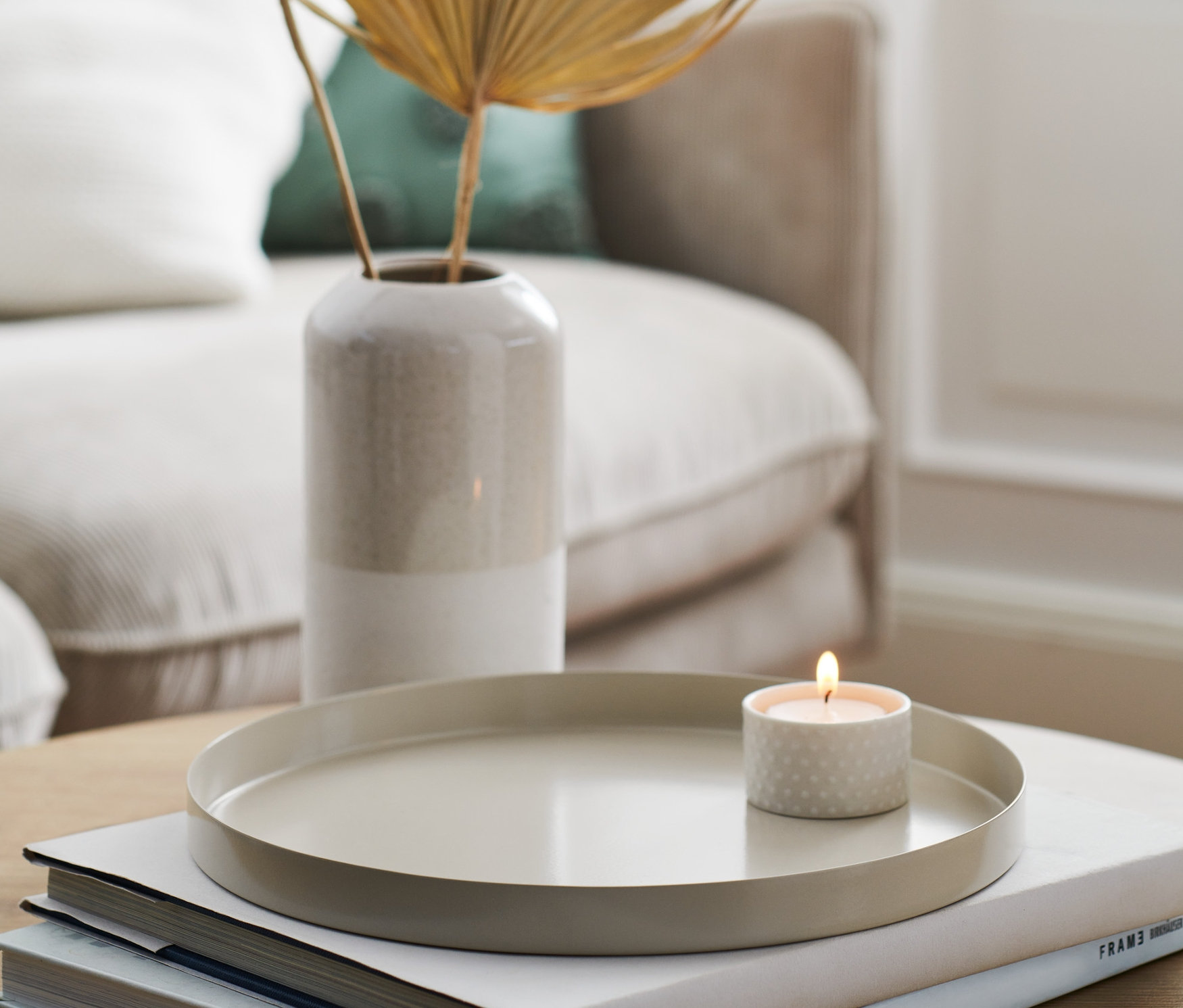 Ein beiges Deko-Tablett mit einer brennenden Kerze auf Büchern neben einer Vase mit Pflanzen.
