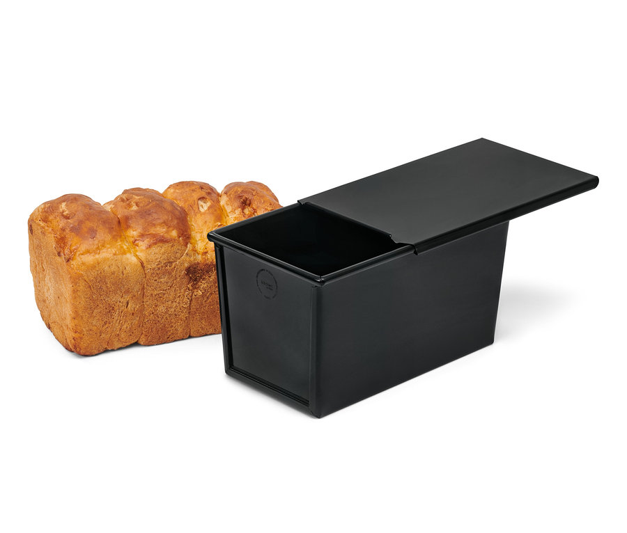 Ein Vollkornbrot liegt neben einer schwarzen Vollkorn-und-Toastbrotbackform.