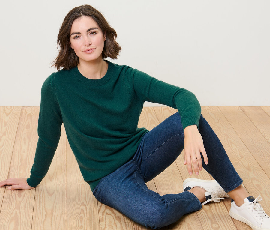 Frau sitzt auf dem Boden und trägt einen grünen Feinstrickpullover mit Cashmere und blaue Jeans.