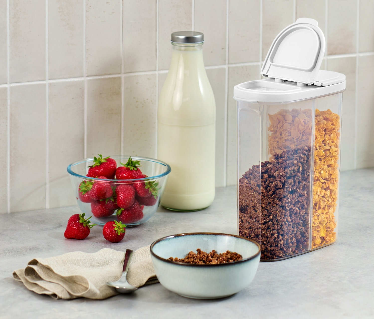 Auf einer Küchentheke stehen eine 2-in-1-Vorratsdose, eine Schüssel Erdbeeren, eine Milchflasche und eine Müslischale.