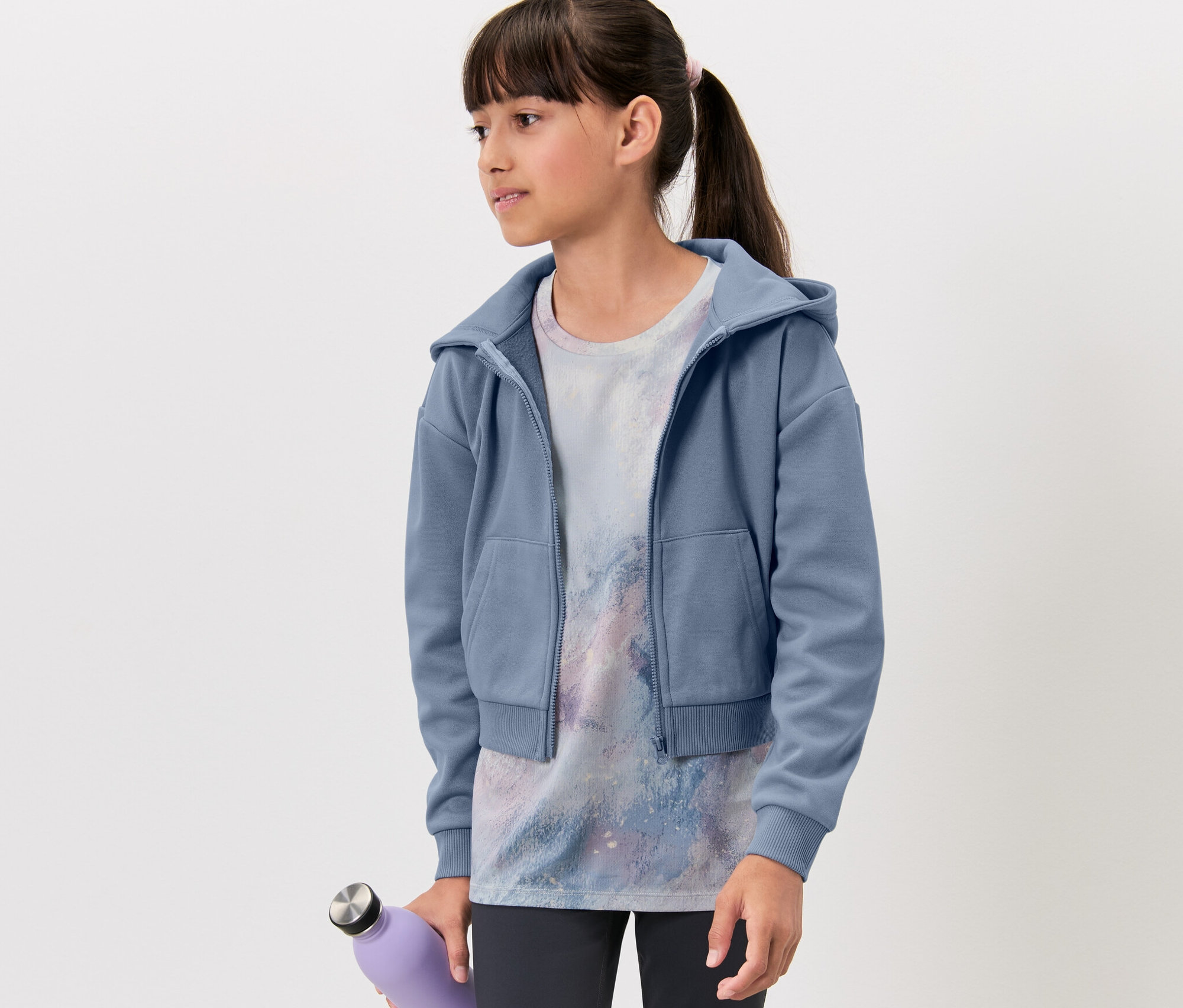 Mädchen trägt eine Kinder-Sportjacke, ein bedrucktes T-Shirt und hält eine lila Flasche.