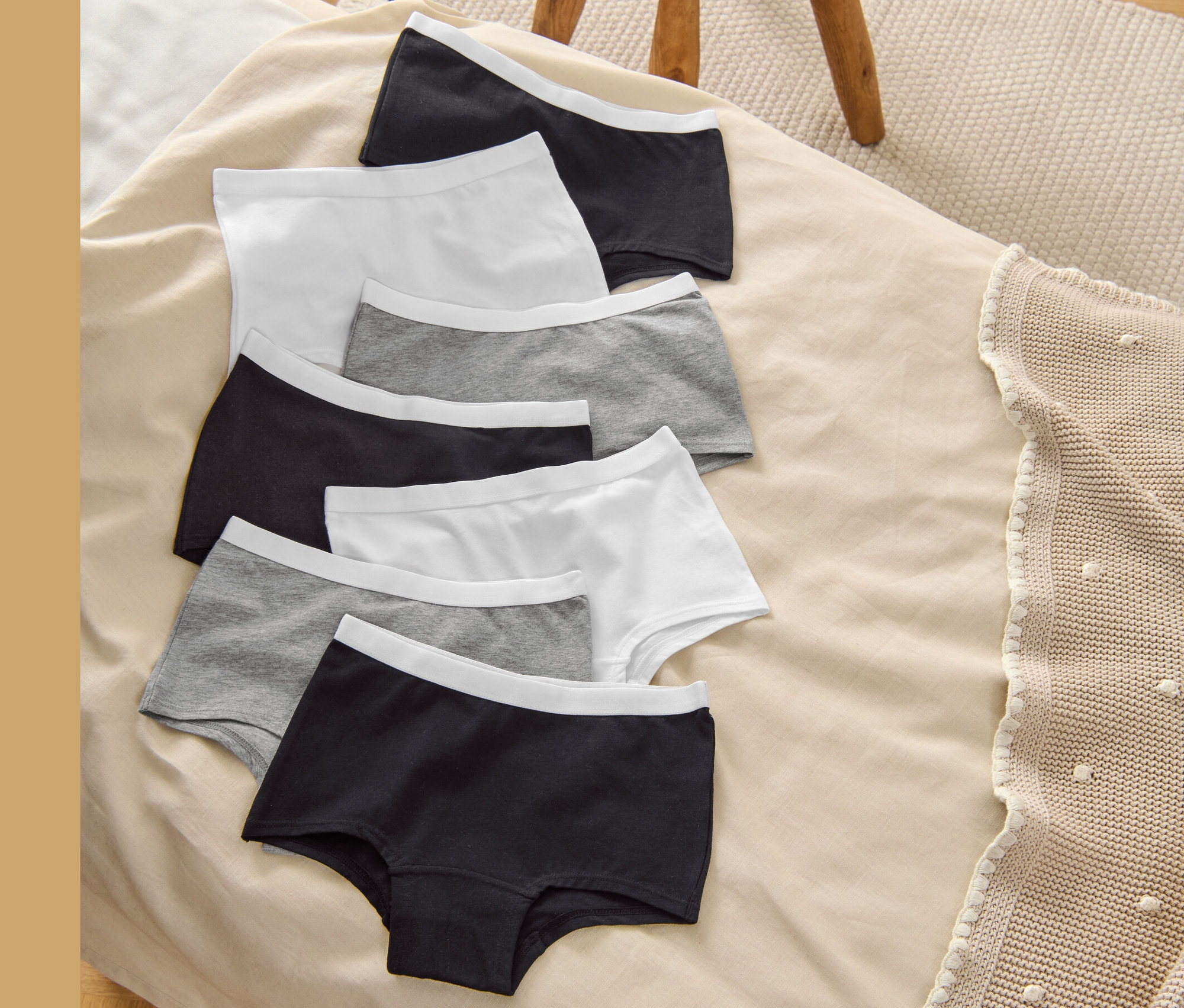 Sieben Kinder-Pantys (7 Kinder-Pantys, grau/weiß/schwarz) liegen auf einer beigen Decke neben einem Holzhocker.