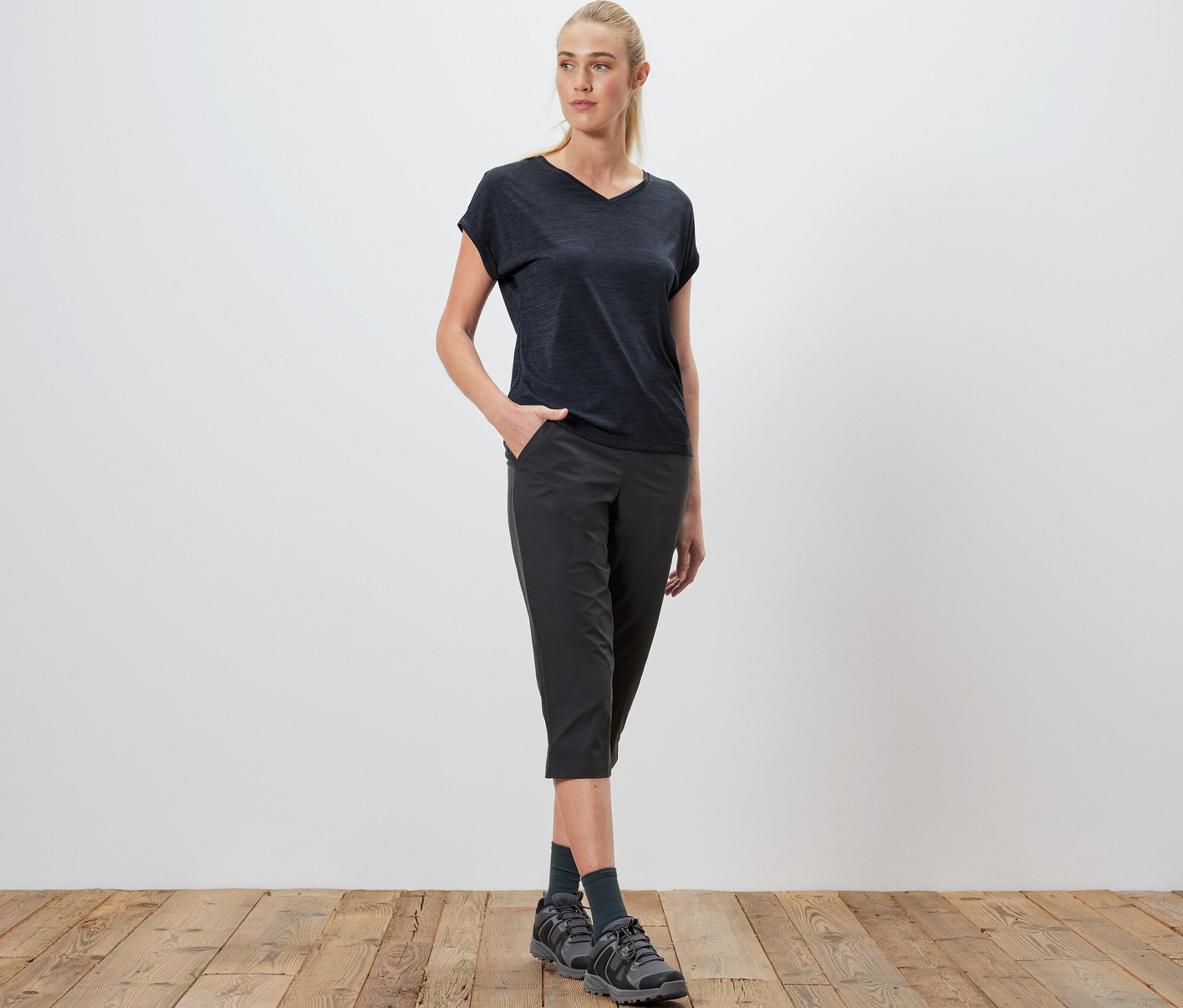 Frau posiert in blauem Funktionsshirt, schwarzen Dreiviertelhosen und Wanderschuhen.