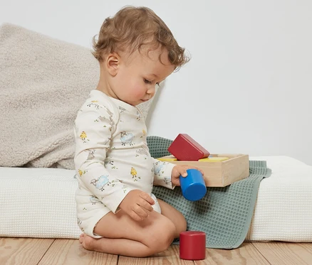 Ein lockiges Kleinkind sitzt auf dem Boden und spielt mit bunten Bauklötzen. Es trägt blaue 3 Baby-Bodys mit Mitwachsfunktion.