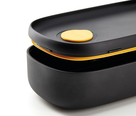 Detailaufnahme eines schwarzen Lunch-Box-Sets mit Besteck, wobei die obere Box leicht geöffnet ist und einen gelben Deckel hat.