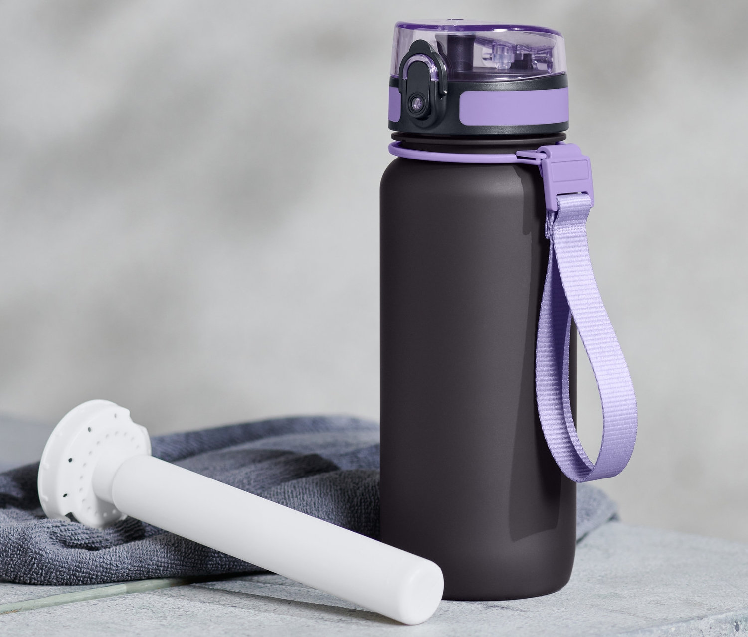 Eine schwarze Trinkflasche mit lila Details und einem weißen Kühlakku liegt auf einer grauen Oberfläche.