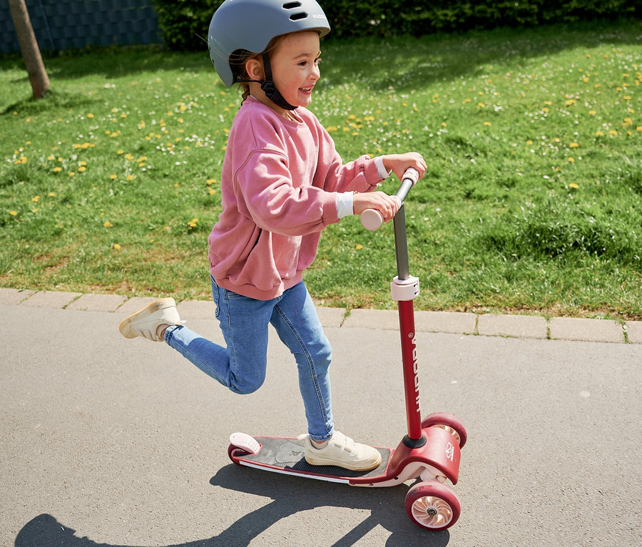 Ein Mädchen fährt mit einem bordeauxroten HUDORA Tri-Scooter und einem grauen Helm auf einem asphaltierten Weg.