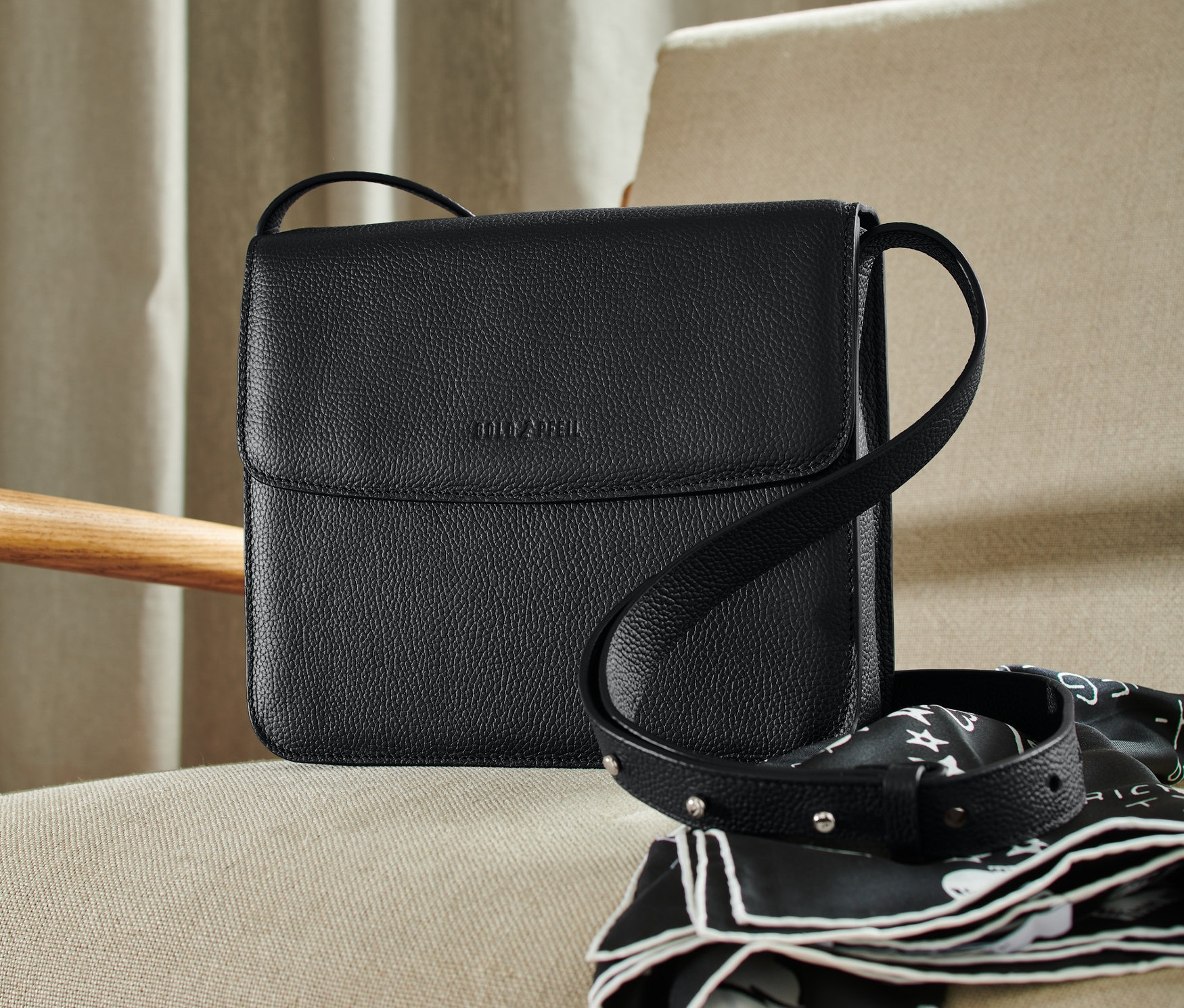 Schwarze Goldpfeil Leder-Umhängetasche liegt auf einem beige Sessel neben einem schwarz-weißen Schal.