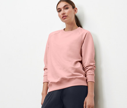 Eine Frau, die vor einer weißen Wand steht, trägt ein roséfarbenes Yoga-Sweatshirt und dunkelblaue Hosen.