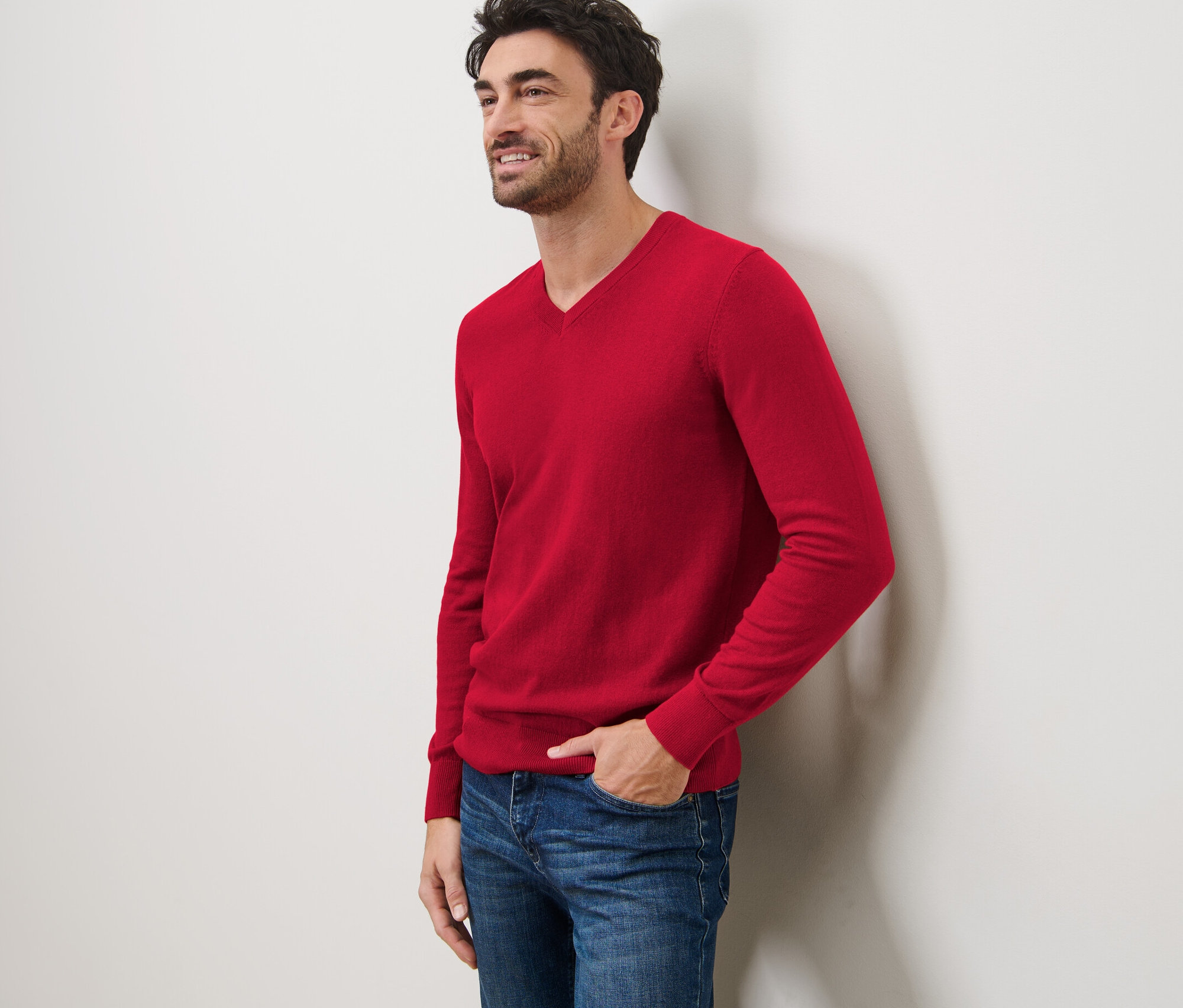 Ein lächelnder Mann lehnt an einer Wand und trägt einen roten Pullover mit V-Ausschnitt und Jeans.