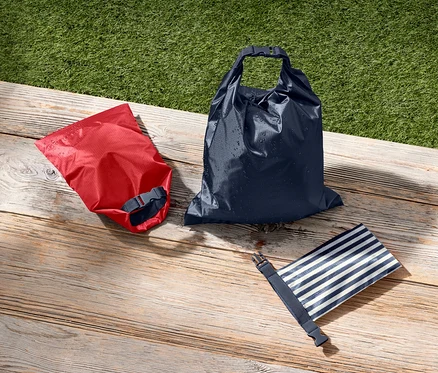 Ein roter, ein dunkelblauer und ein blau-weiss gestreifter wasserdichter Packsack liegen auf einer Holzfläche neben grünem Gras.