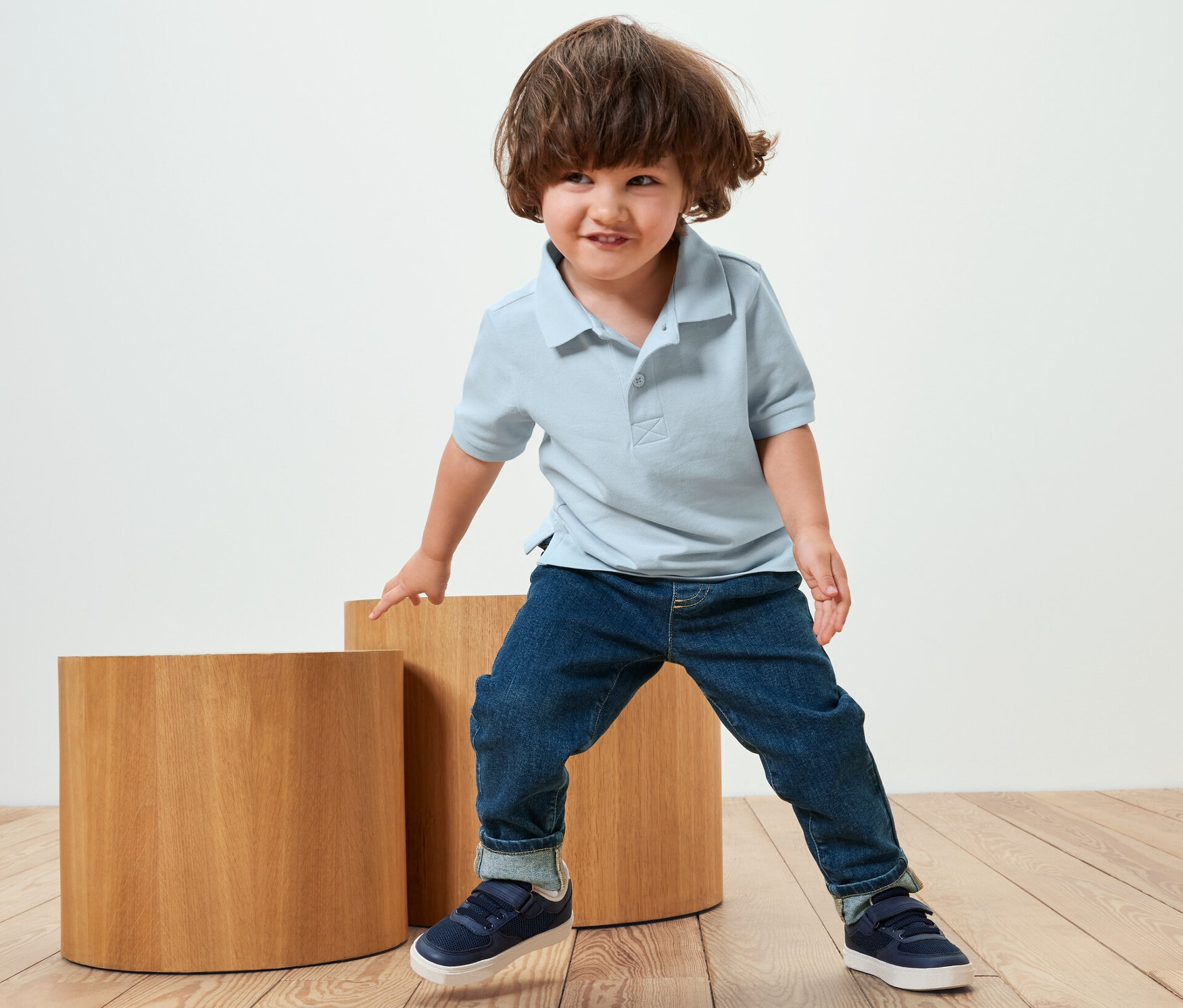 Ein Junge in blauem Poloshirt und dunkelblauer Kinder-Pull-on-Pants – Fit »Luca« steht an zwei Holzsockeln.
