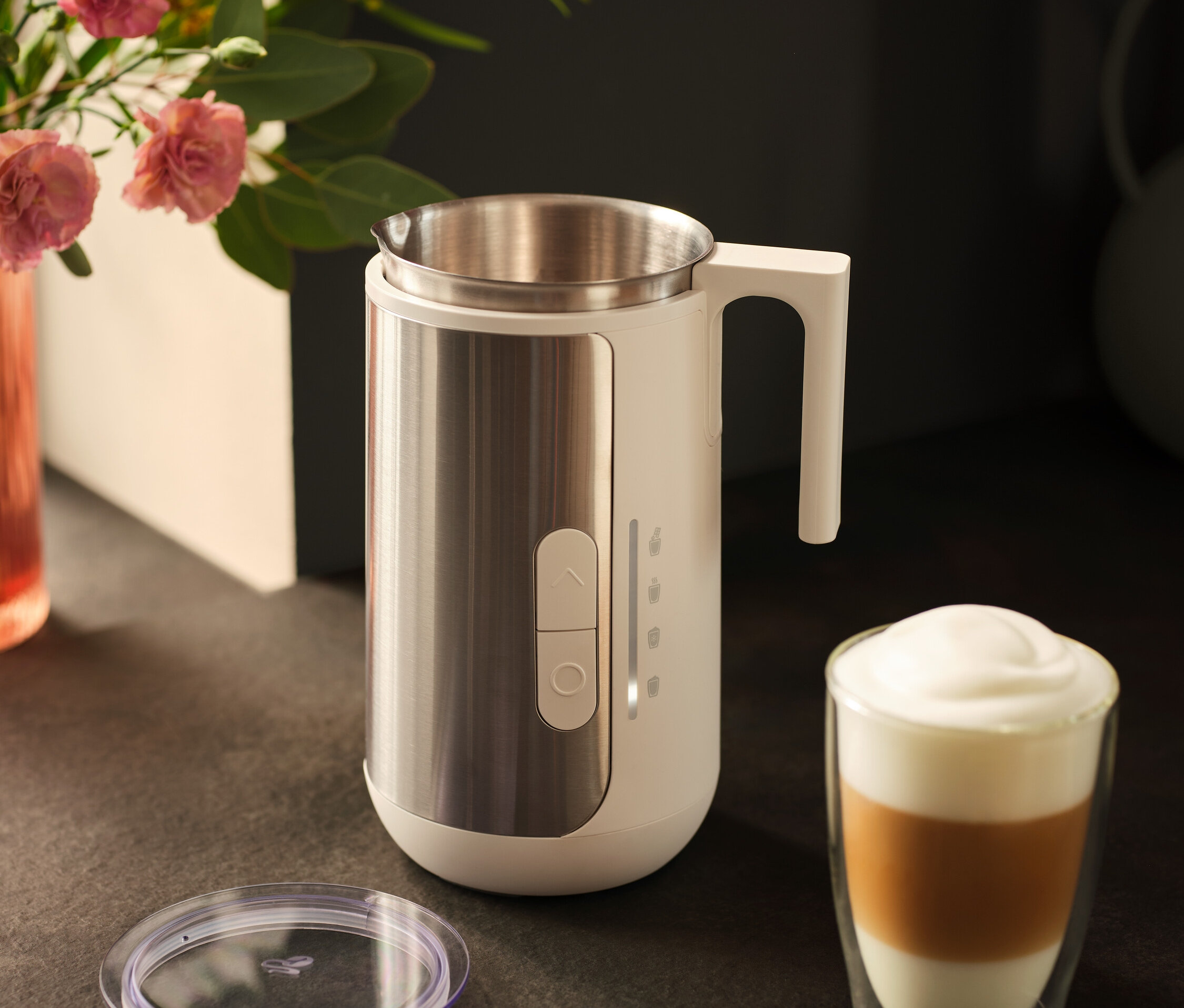 Ein Milchaufschäumer aus Edelstahl steht neben einem Glas Latte Macchiato und einem Deckel auf einer dunklen Arbeitsfläche.
