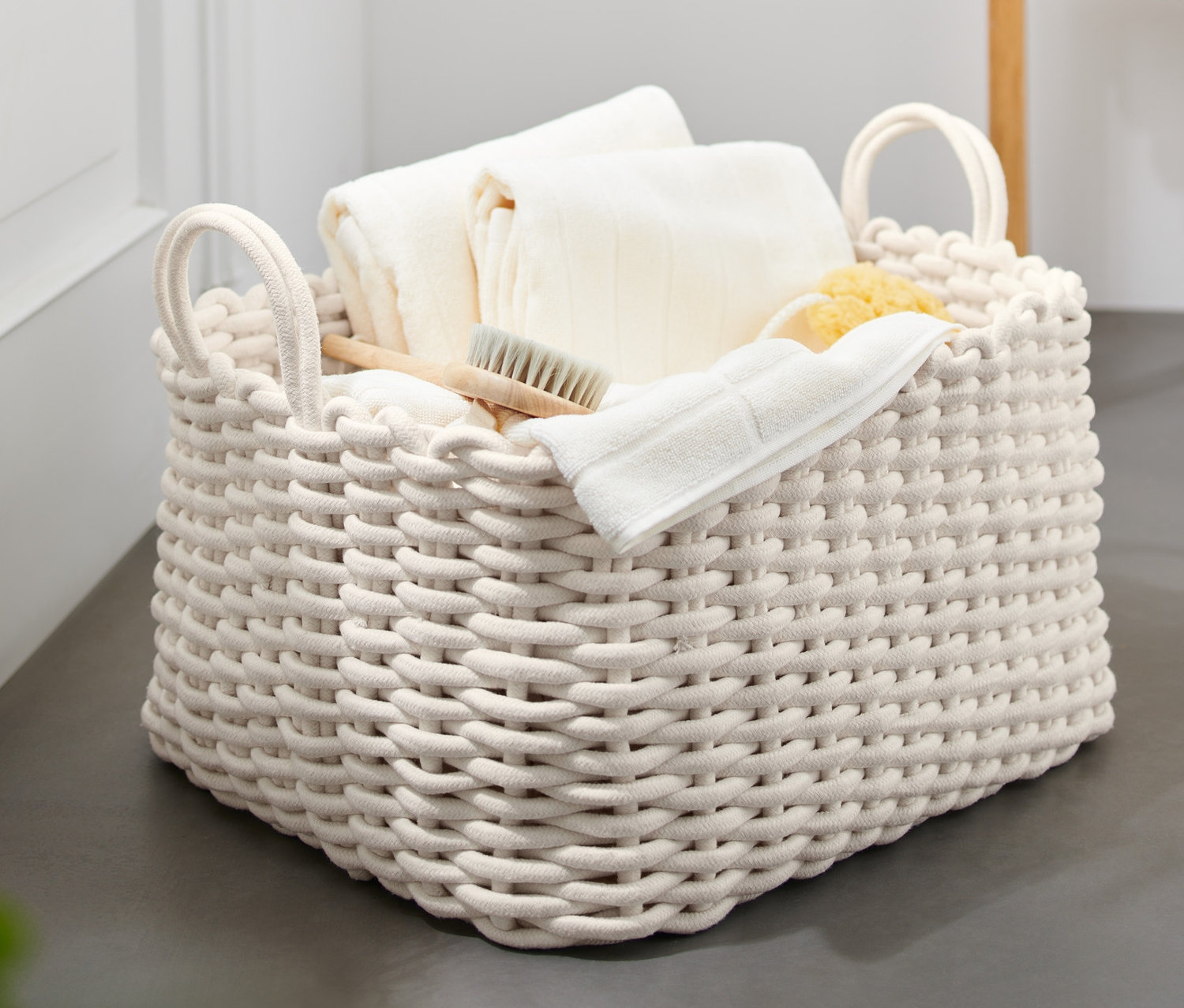 Ein großer, geflochtener Korb mit 2 Henkeln, gefüllt mit weißen Handtüchern, Waschlappen, einer Bürste und einem Schwamm.