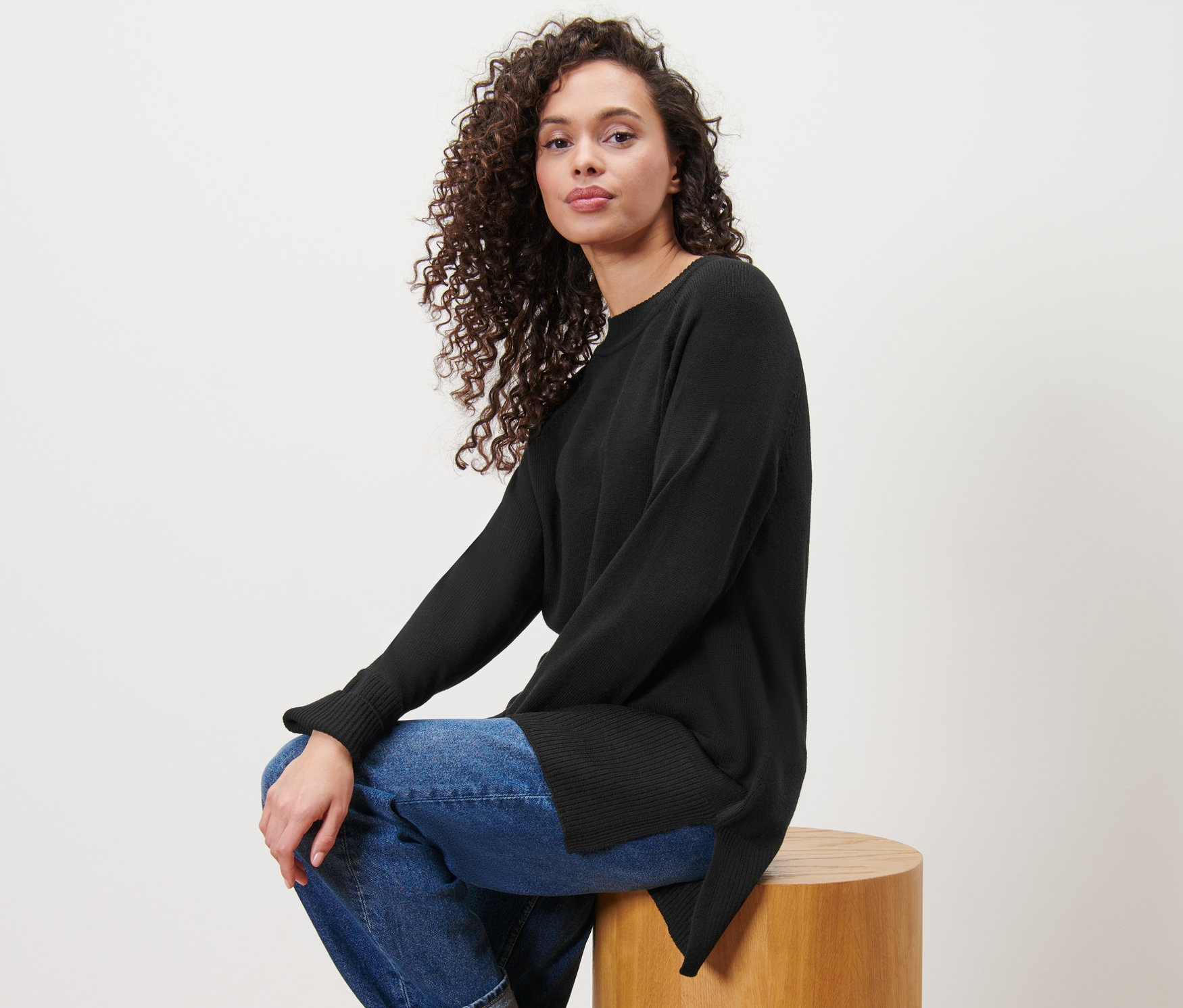 Frau sitzt auf einem Holzhocker und trägt einen langen schwarzen Strickpullover.