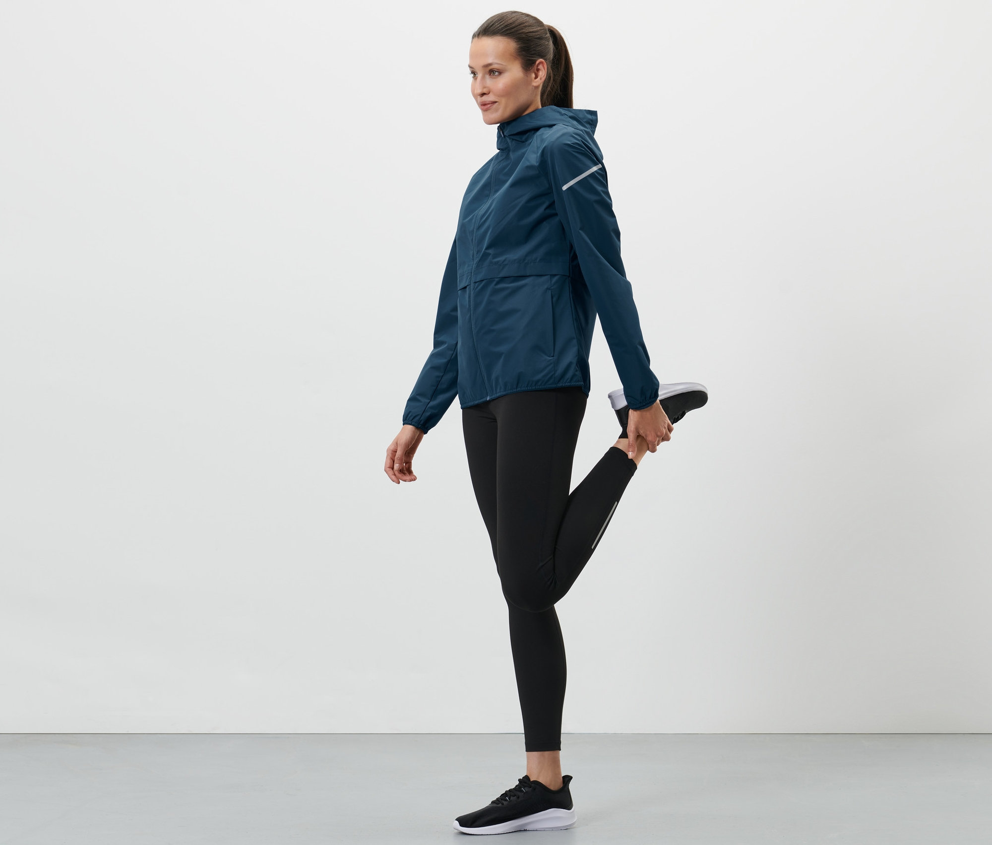 Frau dehnt ihr Bein und trägt eine blaue Windprotection-Laufjacke.