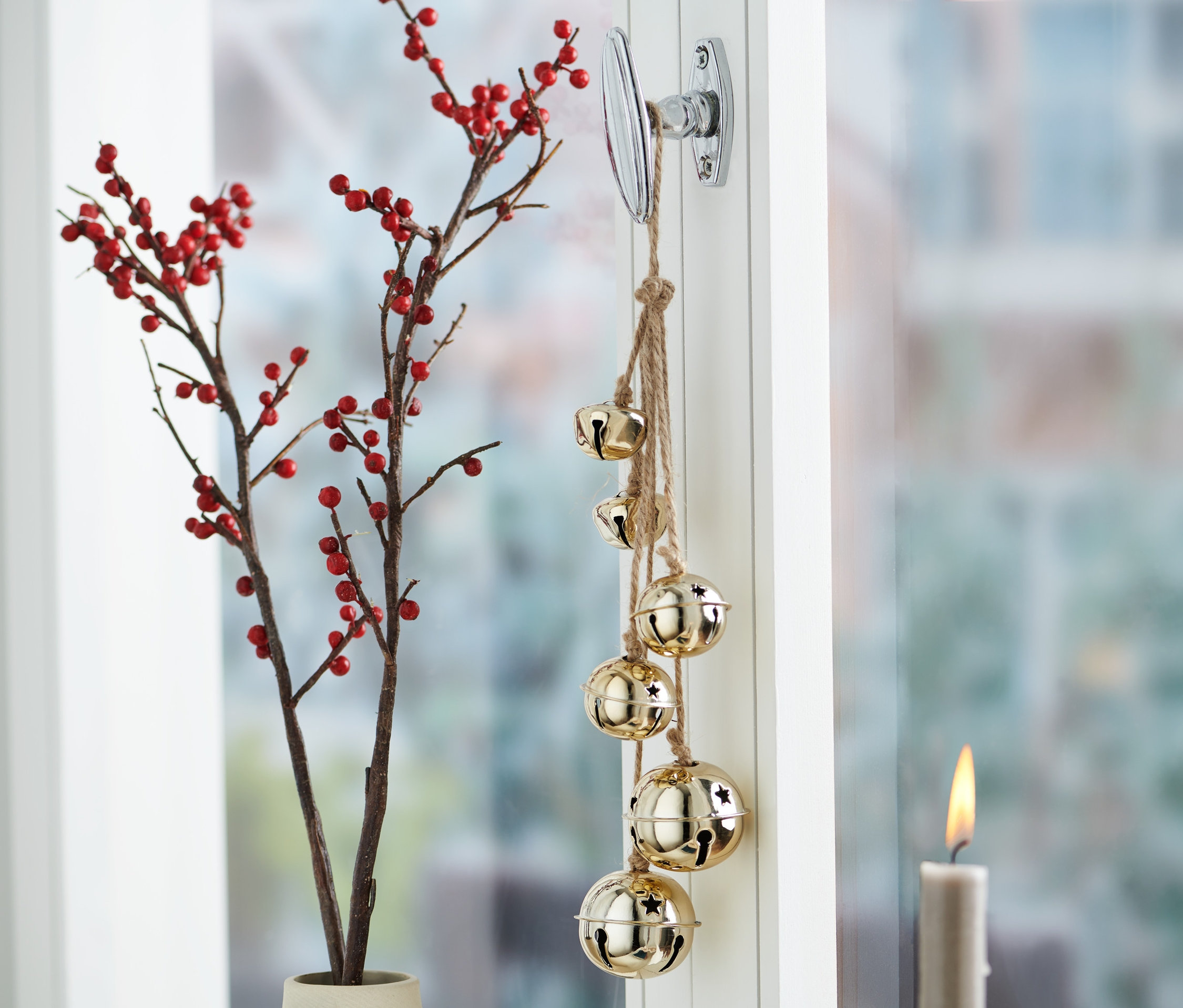 Fensterdekoration mit Glöckchen-Anhängern, Zweigen mit roten Beeren in einer Vase und einer brennenden Kerze.