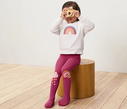 Mädchen sitzt auf einem Holzhocker und schaut durch eine Holzkamera. Sie trägt eine 3 Paar Kinder-Strumpfhosen, rosa.