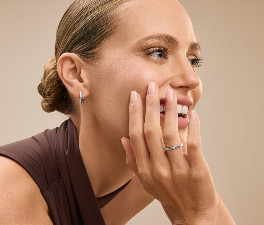 Porträt einer Frau mit zum Dutt hochgesteckten Haaren, die lächelt und ihr Gesicht mit einer Hand berührt, an der sie einen silbernen Ring trägt.
