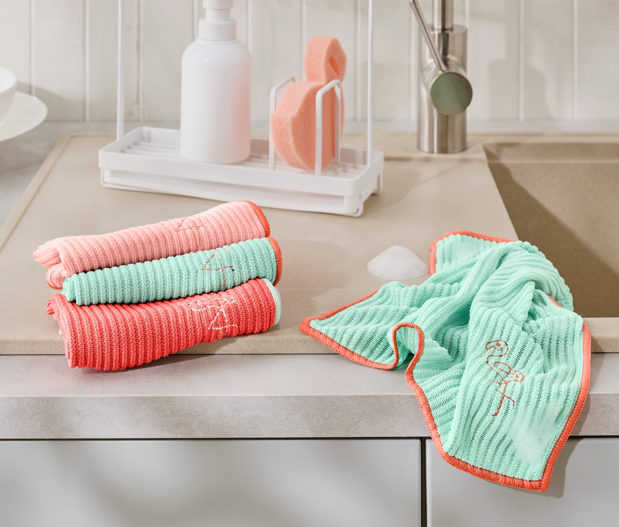 Auf einer Küchentheke liegen gefaltet und ausgebreitet 4 Universal-Reinigungstücher in Koralle, Rosa und Türkis mit Flamingostickerei.