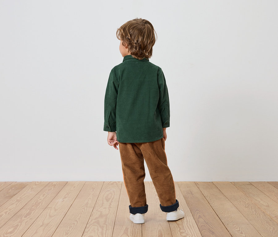Ein Junge in einem grünen Hemd und braunen Hosen steht mit dem Rücken zum Betrachter auf einem Holzboden.