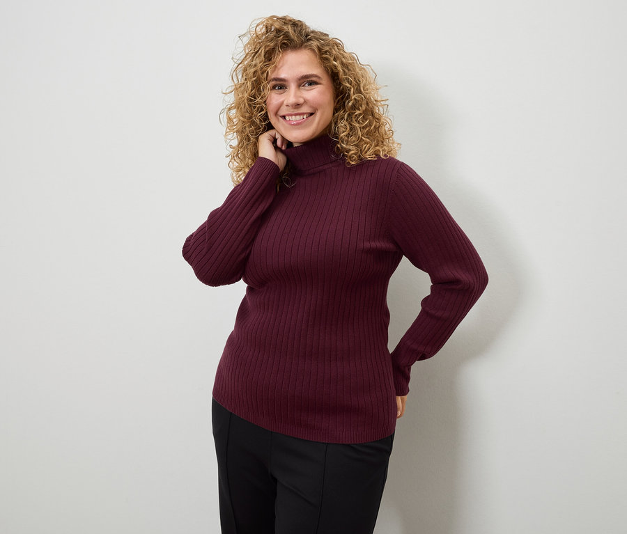 Eine Frau mit lockigen Haaren posiert in einem burgunderroten Rollkragenpullover und einer schwarzen Hose.